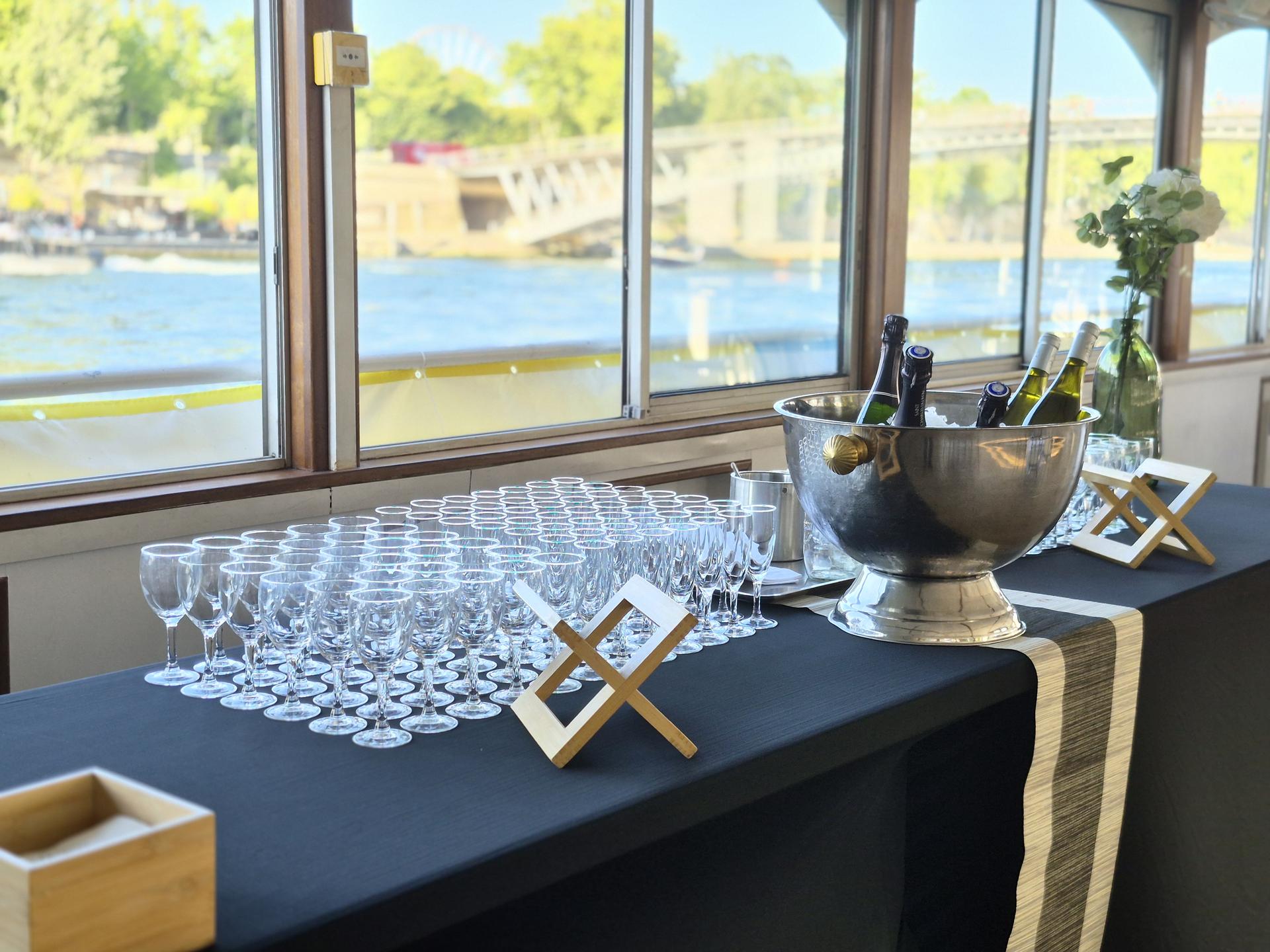 Buffet de verres et seau à champagne devant les baies d'un bateau, la Seine au soleil