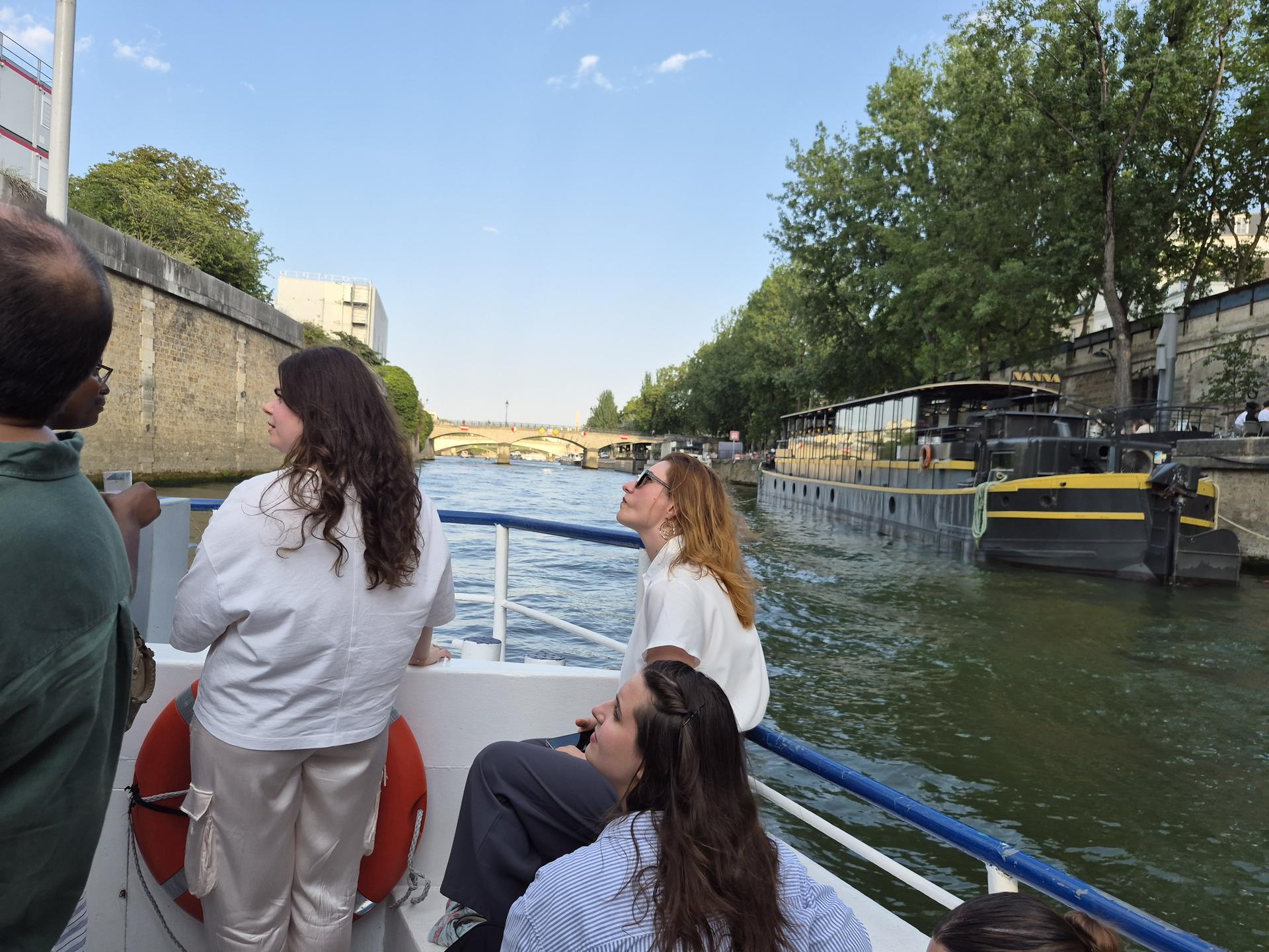 Invités à l'avant d'un bateau sur la Seine, péniche noire et jaune amarrée, ciel bleu